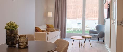 Un salon moderne avec une table à manger ronde, un canapé et un fauteuil. Il y a un balcon avec vue sur un immeuble en briques.