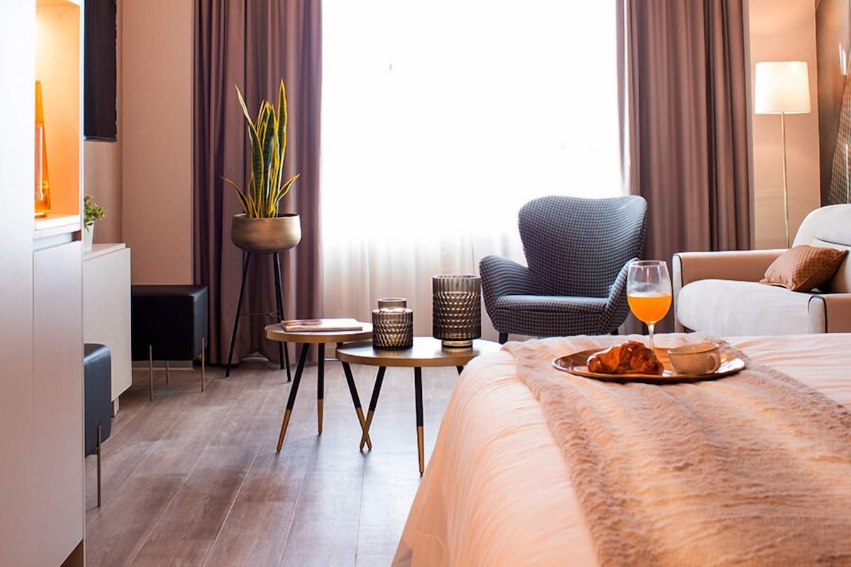 Una habitación de hotel moderna con una cama grande, un escritorio, una silla, una planta en maceta y un ventanal con cortinas.