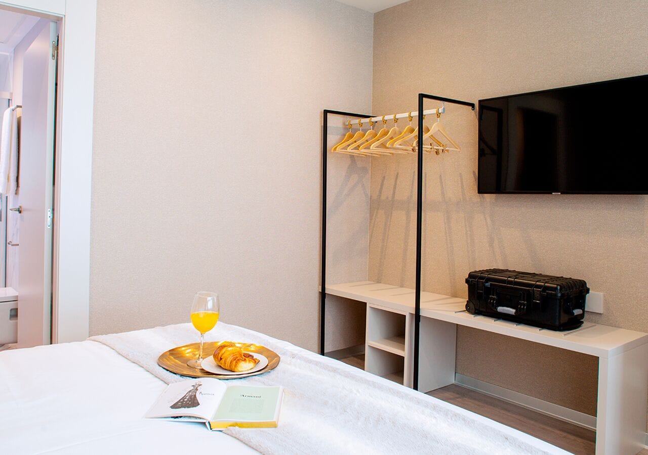 A hotel room with a bed, a breakfast tray with croissants and orange juice, a suitcase, and a TV mounted on the wall.