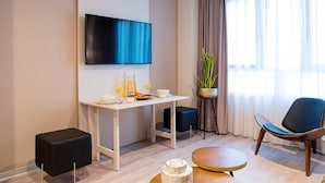 Une chambre d’hôtel moderne équipée d’un téléviseur à écran plat, d’une table à manger avec des produits pour le petit-déjeuner, d’une plante en pot, d’un fauteuil confortable et d’un petit pouf noir.