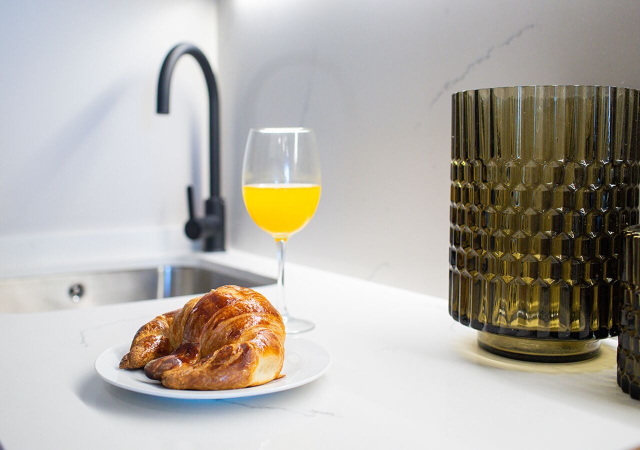 En croissant på en tallrik, ett glas apelsinjuice och en vas med intressant yta på en vit bänkskiva.