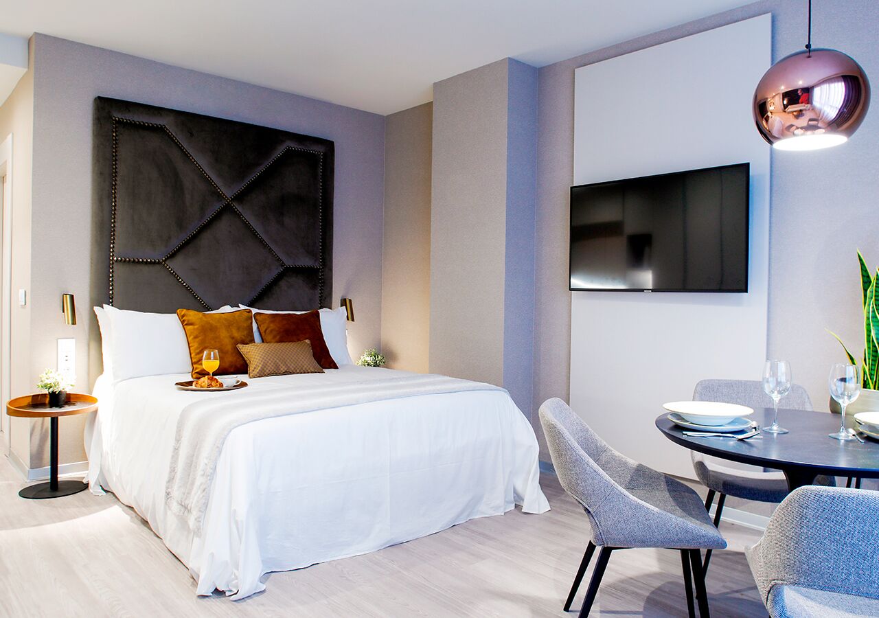 Ein modernes Hotelzimmer mit einem großen Bett, einem flachbildfernseher an der Wand, einem runden Esstisch mit Stühlen und einem kleinen Nachttisch mit Lampe.