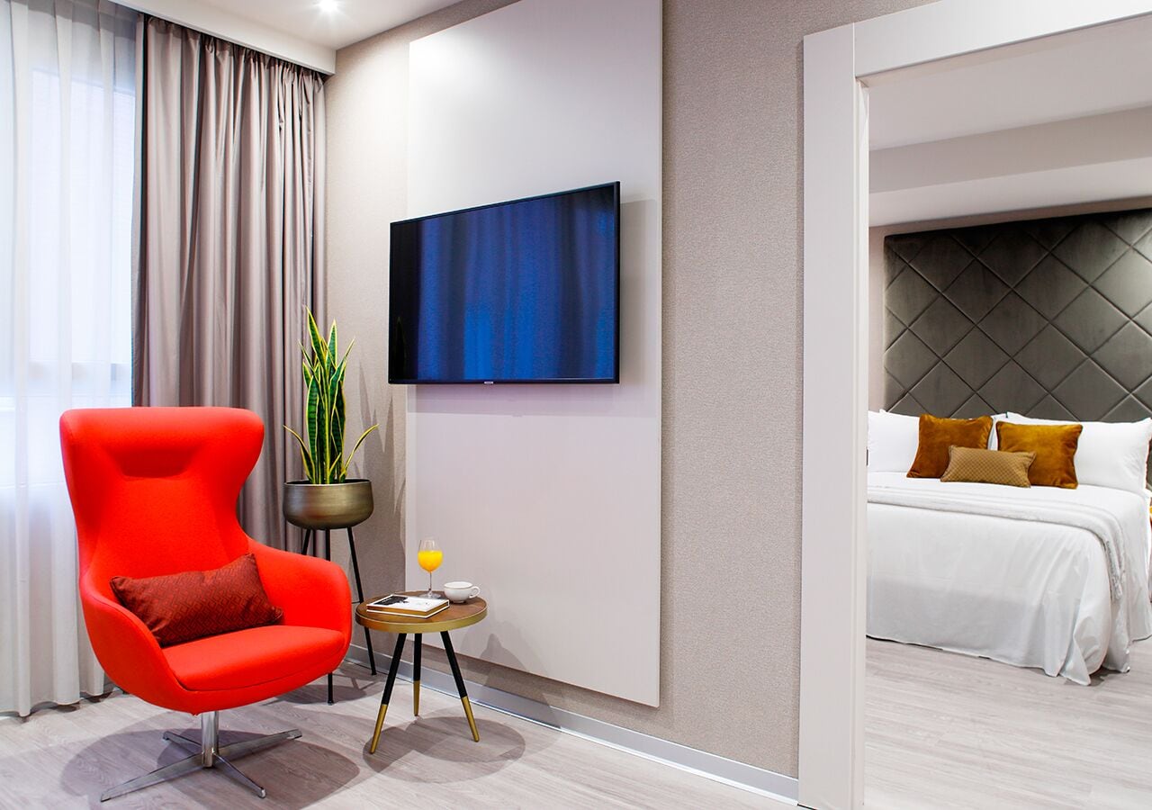 A modern hotel room with a red armchair, a small table with a glass of orange juice, a potted plant, a flat-screen TV, and a bed with white bedding and brown pillows.