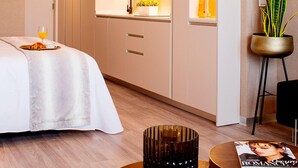 Une chambre d’hôtel moderne comprenant un lit, une table ronde avec un magazine, une petite plante et un coin cuisine avec un évier et des placards.