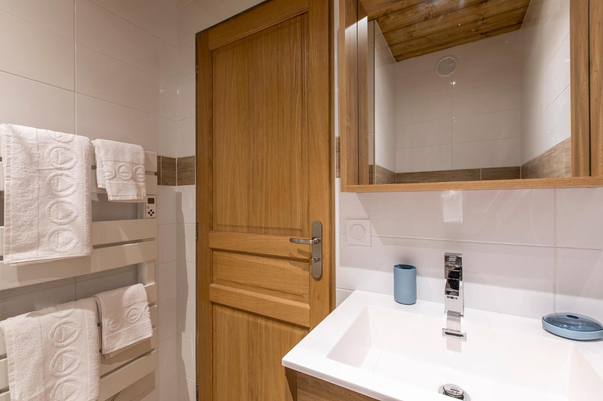 A bathroom with a wooden door, white tiled walls, a white sink, and a mirror above it.