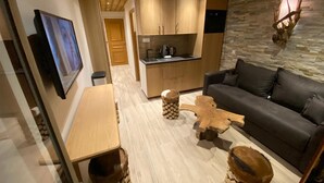 A modern living room with a wooden coffee table, a black sofa, and a kitchen area with wooden cabinets and a stone wall. - Résidence La Forêt Du Praz (Courchevel)