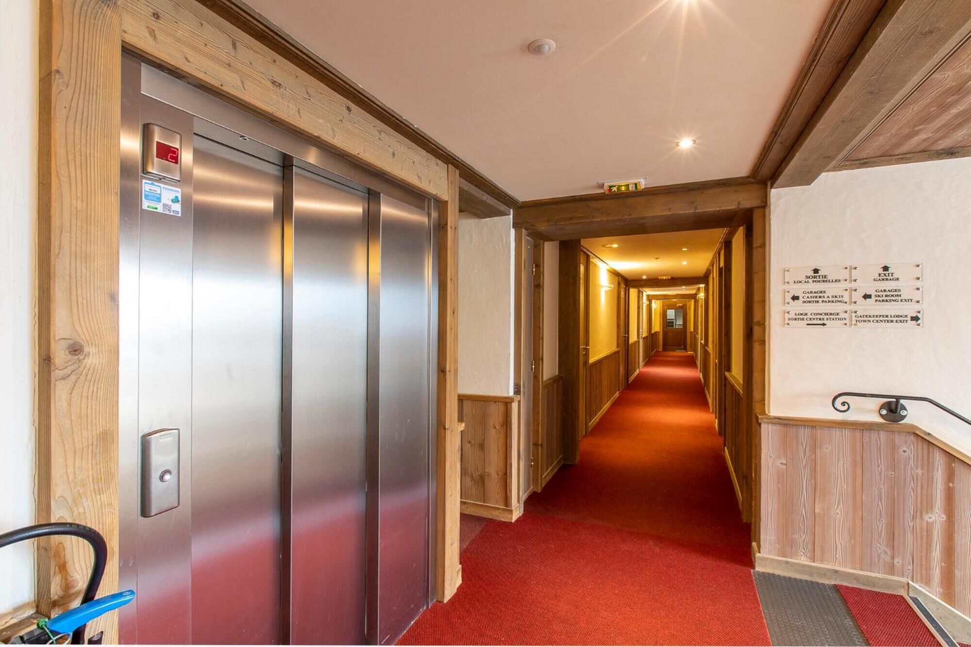Un couloir d’hôtel avec un ascenseur, une moquette rouge, des lambris en bois et des panneaux de signalisation.