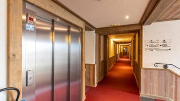 Un couloir d’hôtel avec un ascenseur, une moquette rouge, des lambris en bois et des panneaux de signalisation.