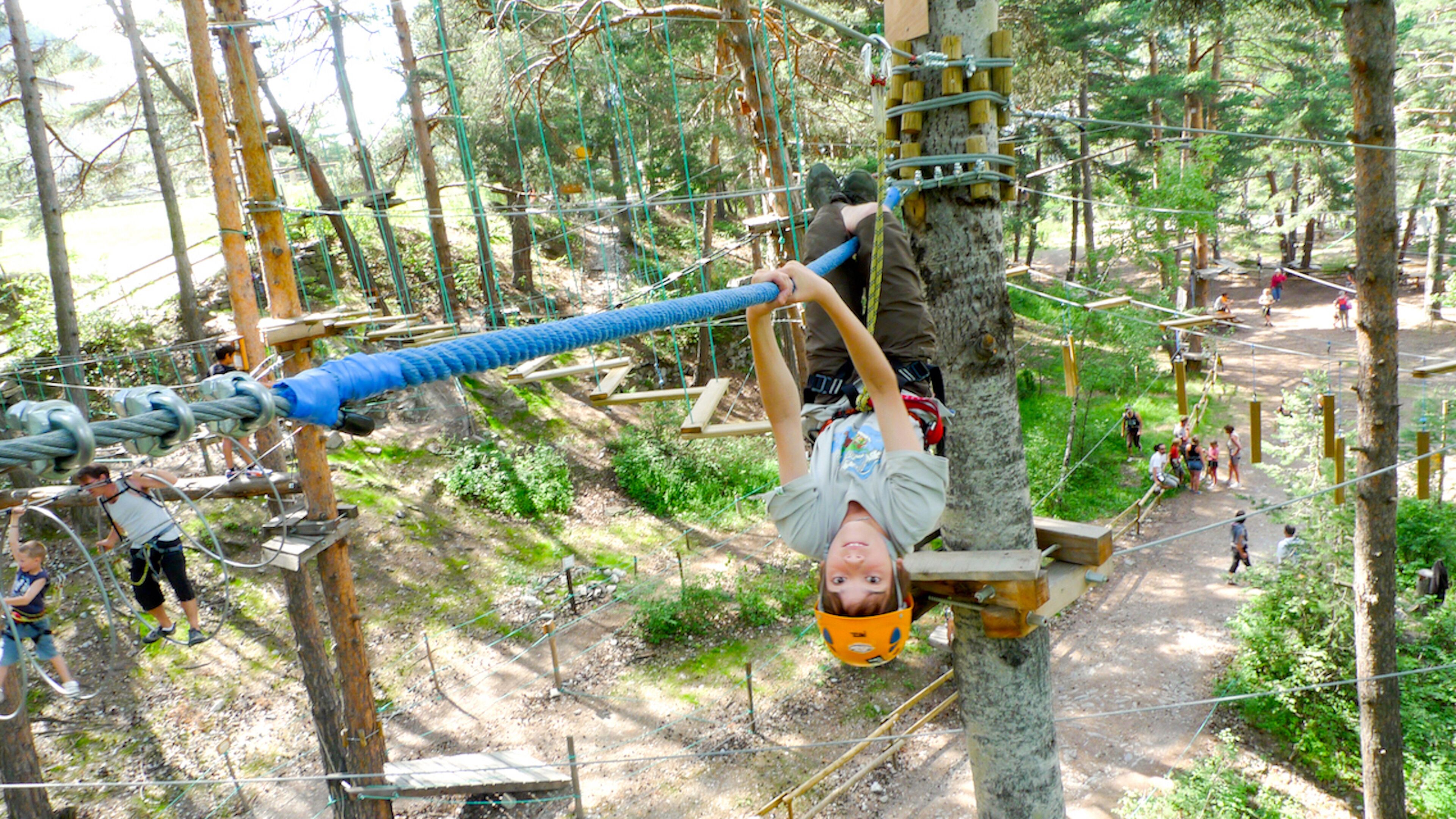 A person suspended on a rope course in a forested area with multiple participants and safety equipment visible.