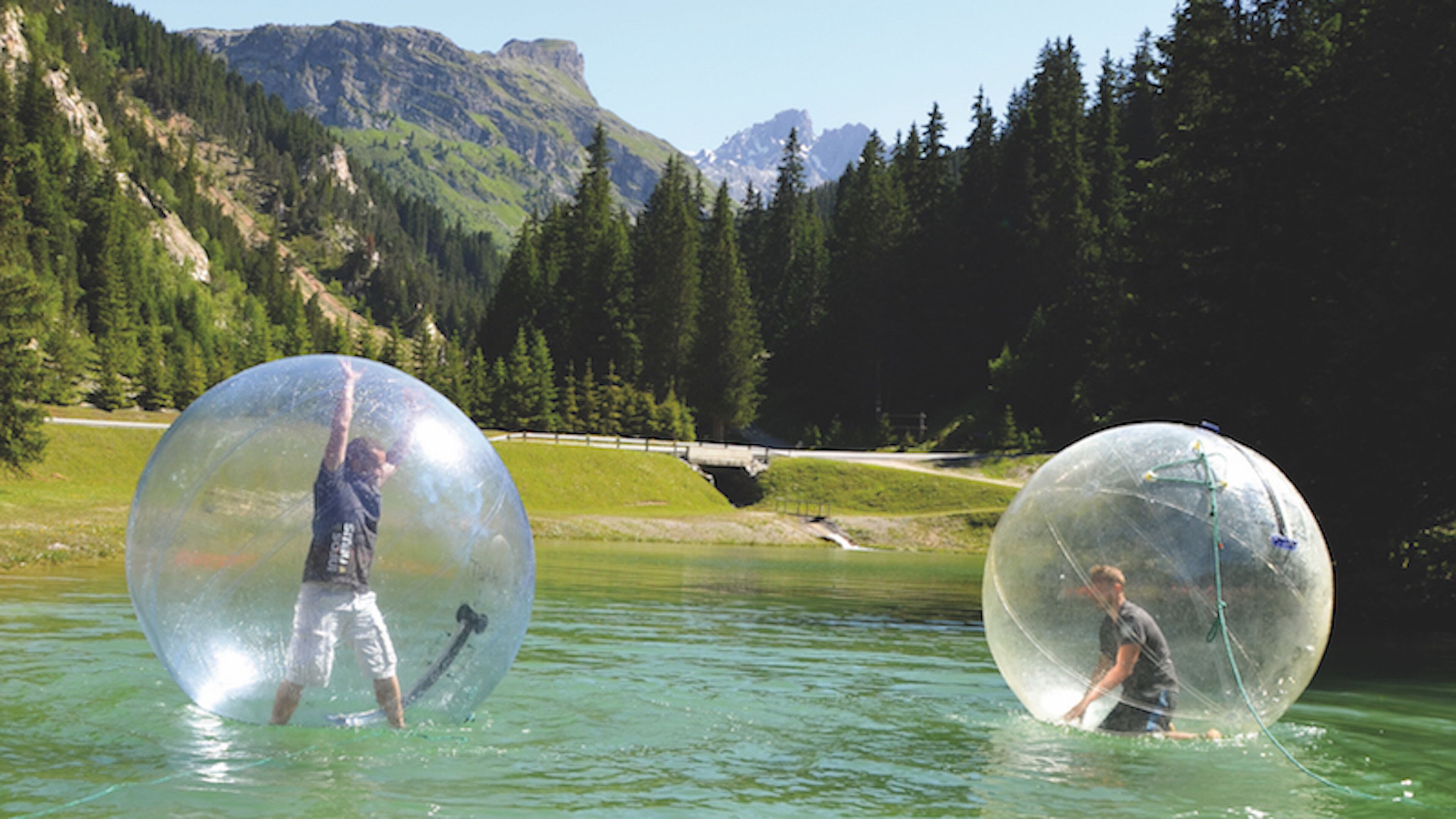 Twee personen in doorzichtige bolvormige capsules, drijvend op het water met bergen op de achtergrond.