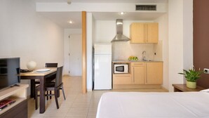 A compact hotel room with a bed, a TV, a dining area with a table and chairs, a kitchenette with a microwave, and a small plant. - SG Marina 54 Apartments (Castelldefels)