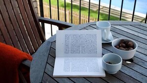 A wooden table with a book, two white cups, and a bowl of nuts on a balcony overlooking a swimming pool. - SG Marina 54 Apartments (Castelldefels)