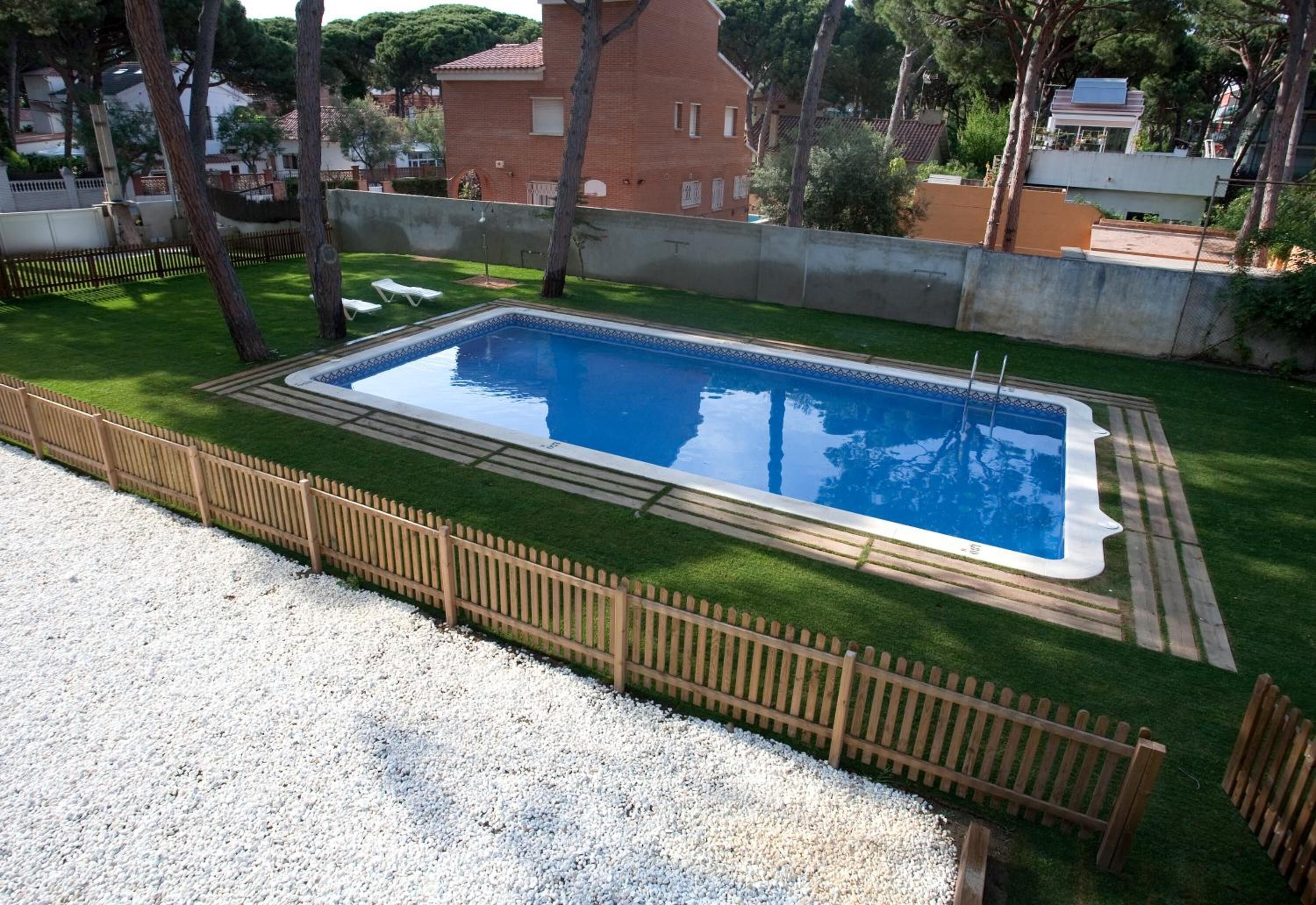 Una piscina rectangular con una terraza de madera alrededor, ubicada en un área cubierta de césped con un camino de grava que lleva a la piscina.