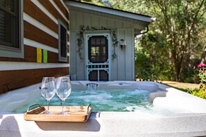 A hot tub with two glasses and a tray, a wooden house with a white door, and a garden with trees and flowers.