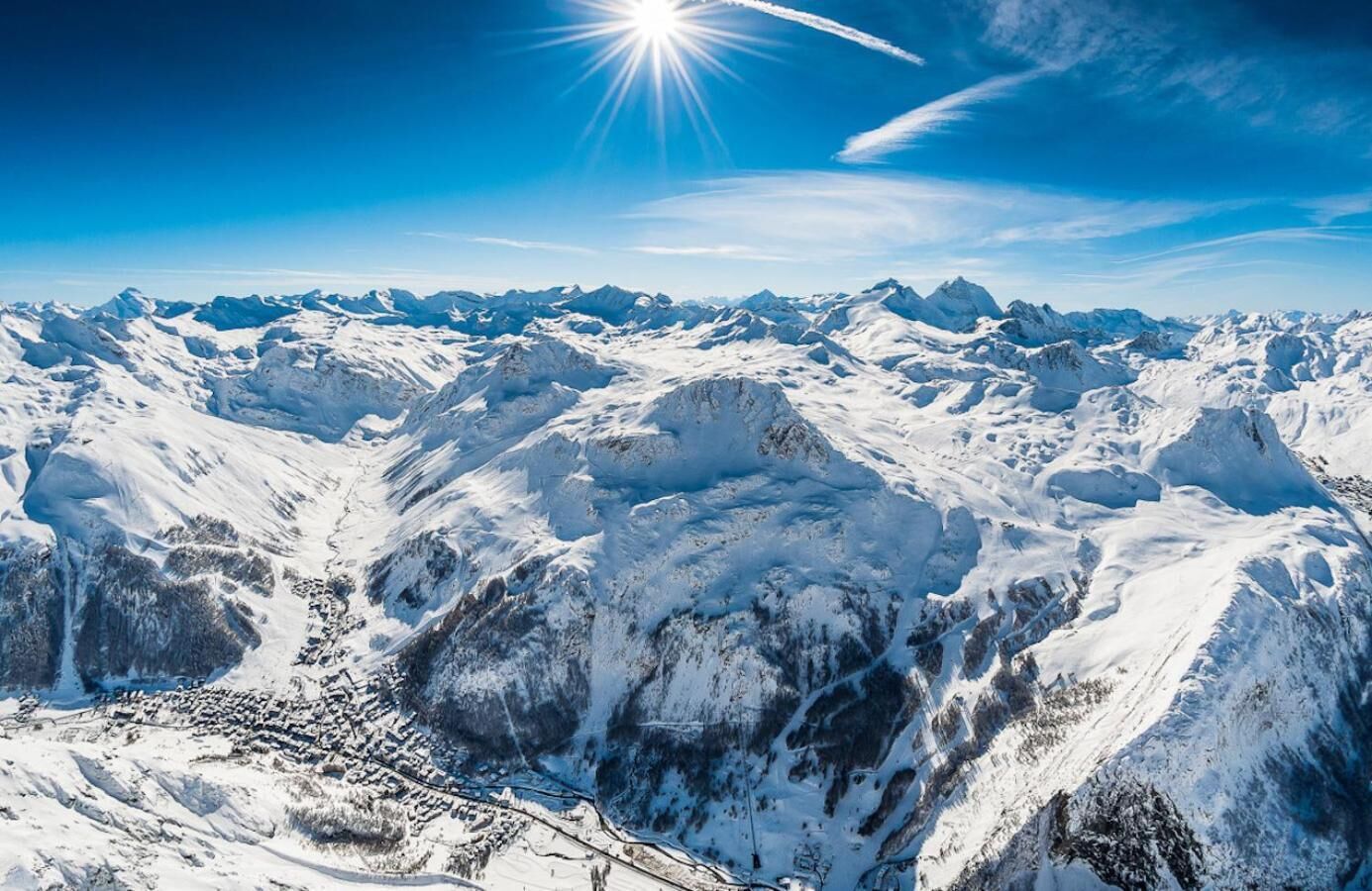A panoramic view of a snow-covered mountain range with a clear sky and a bright sun.