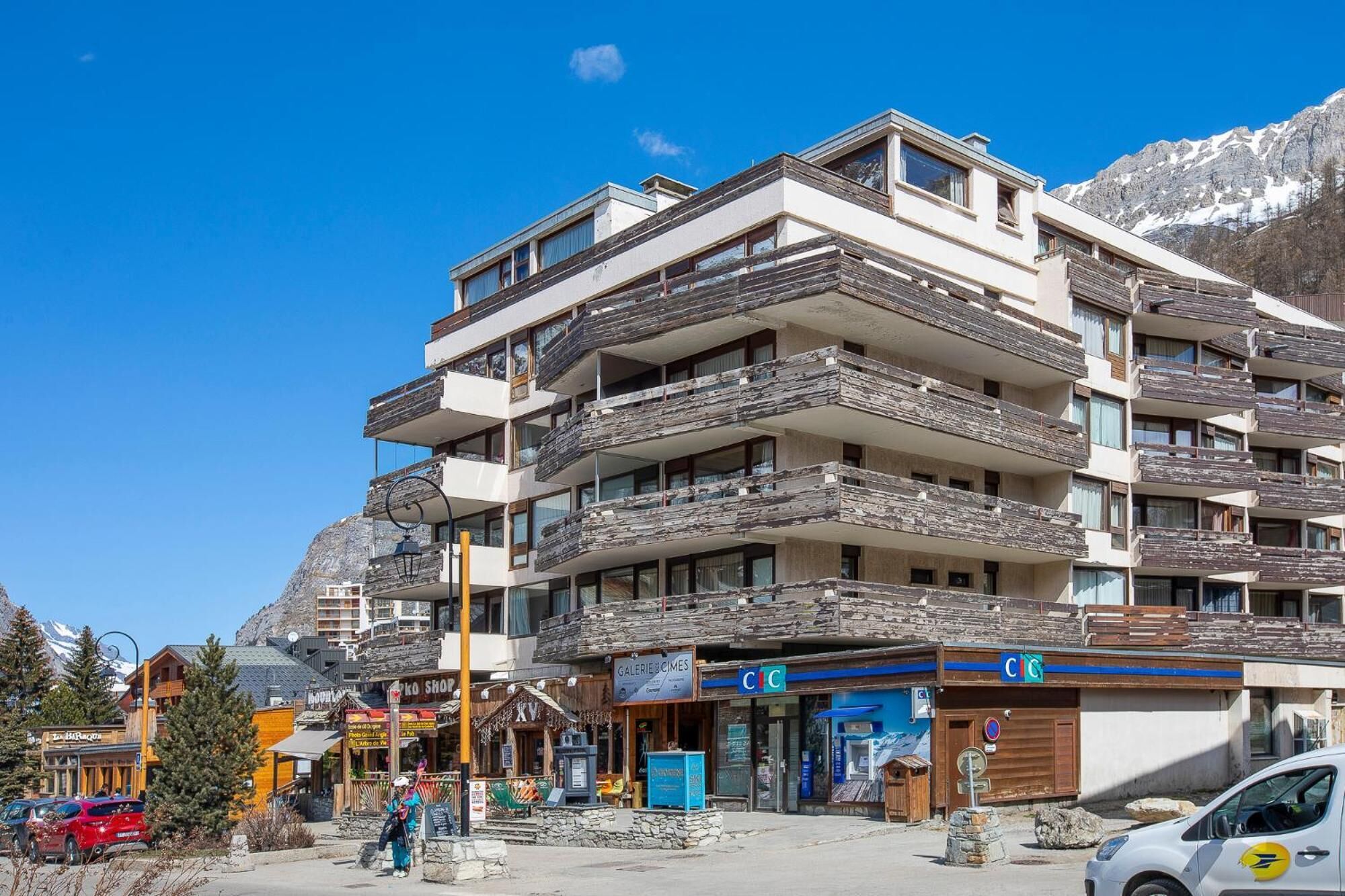 A multi-story hotel with balconies, shops on the ground floor, and a mountainous backdrop.