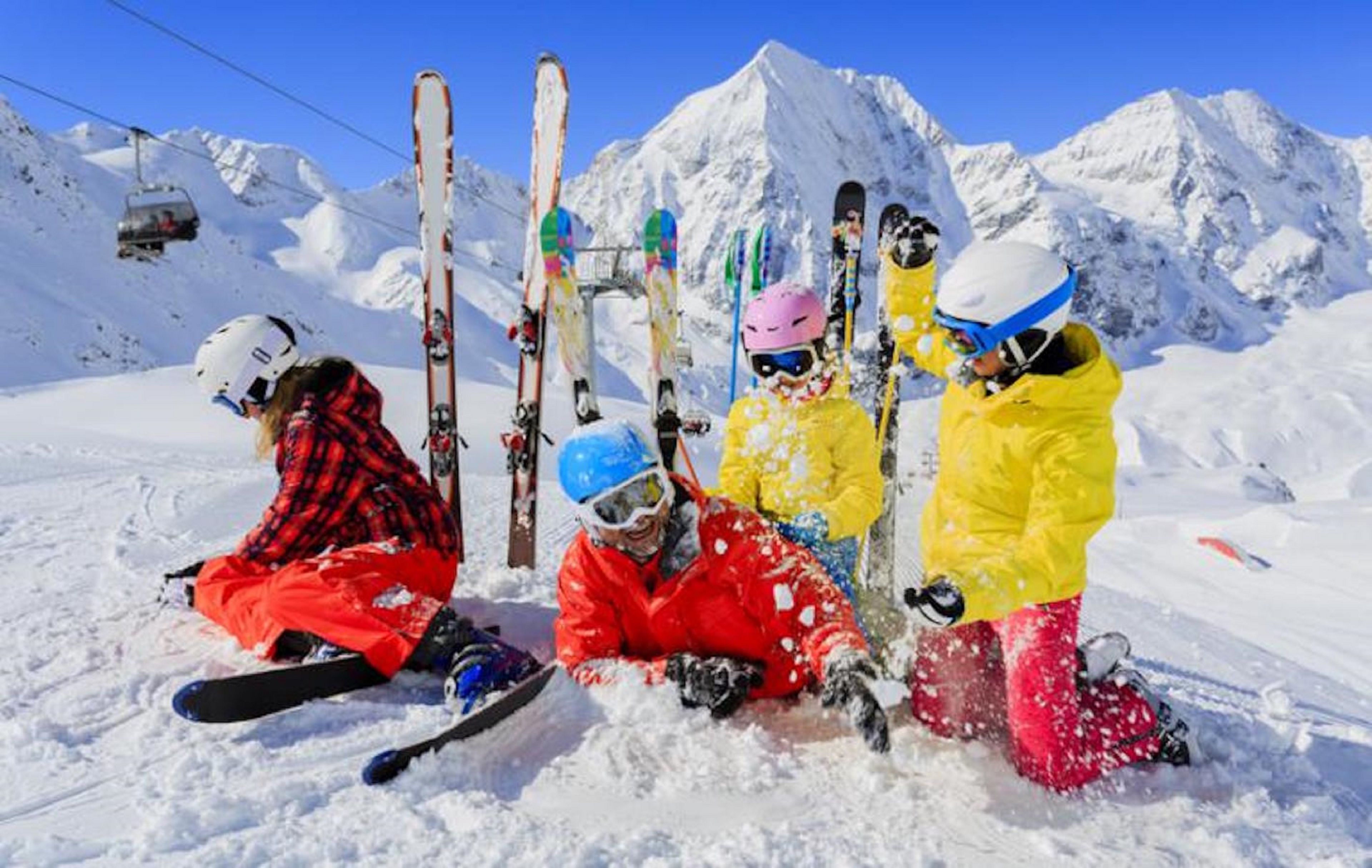 Skiers sitting on the snow with skis leaning against a pole, a ski lift in the background, and snow-covered mountains.
