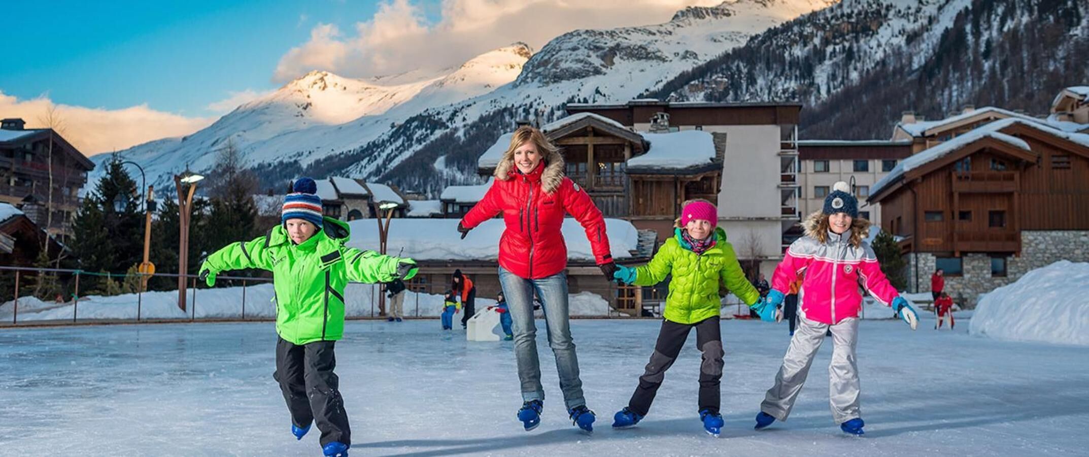 A group of people ice skating in a snowy outdoor area with wooden buildings in the background.