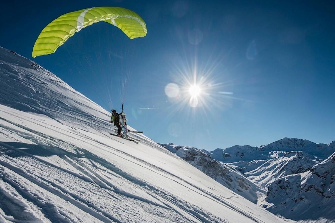 A person skiing down a snowy mountain slope with a parachute, under a clear blue sky.