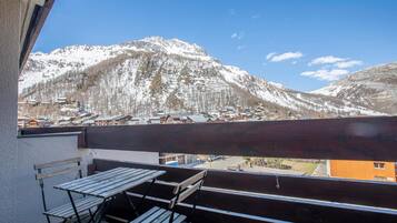 Un balcon doté de mobilier en bois, offrant une vue sur un paysage montagneux avec des sommets enneigés et un petit village en contrebas.
