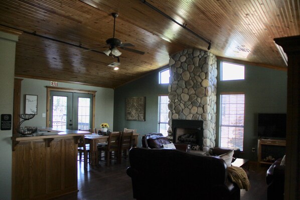 A spacious living area with a stone fireplace, wooden ceiling, and a dining area with a bar.