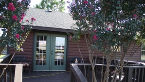 A log cabin with a green door and windows, a deck with a bench, and a tree with pink flowers. - The Outdoorsman at Homes on Holiday (Ardmore)
