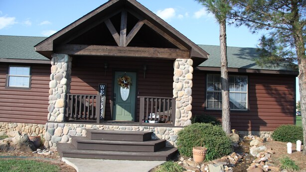 A wooden cabin with a stone facade, a front porch, and a small garden area with rocks and plants. - The Outdoorsman at Homes on Holiday (Ardmore)