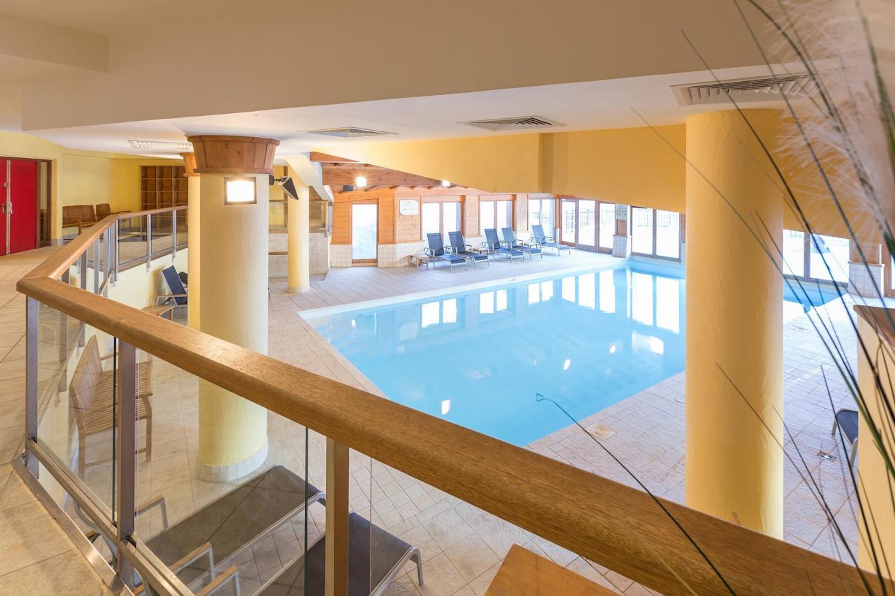 Een binnenzwembad met glazen balustrade, houten zuilen en zonnige kamers op de achtergrond.
