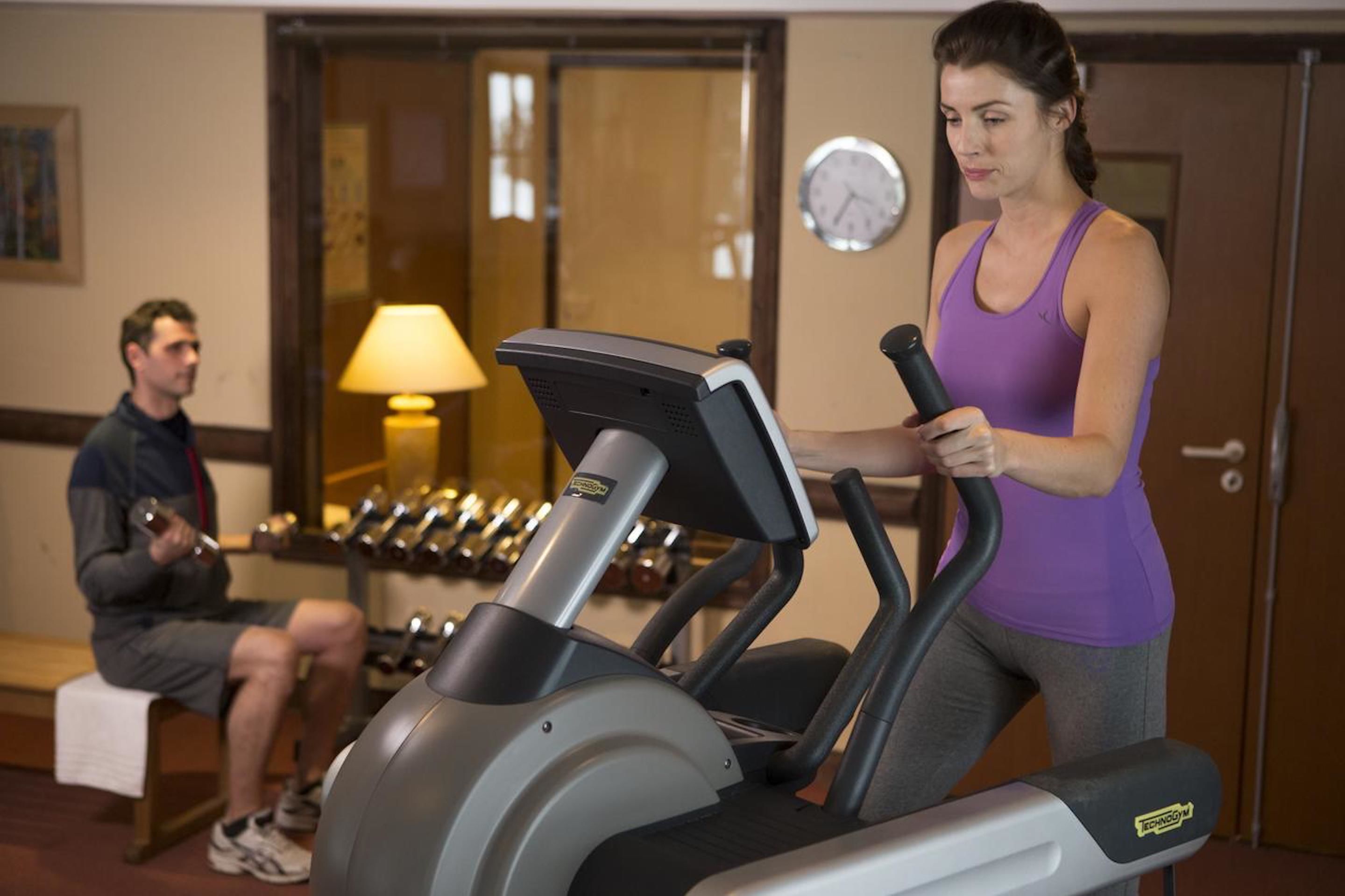 Un gimnasio con cintas de correr, un hombre usando mancuernas y una mujer en una cinta de correr.