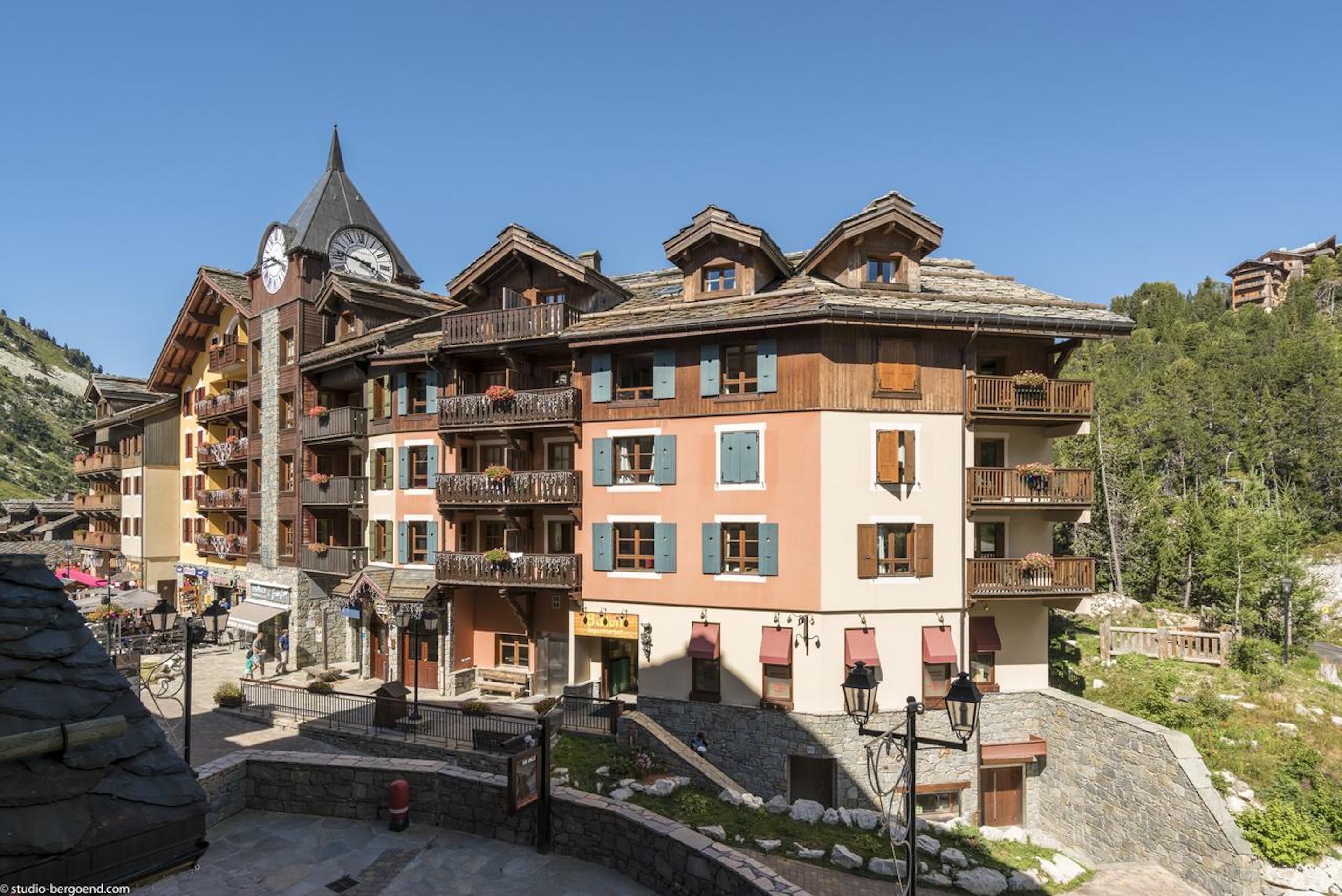 A traditional chalet-style building with multiple balconies and a clock tower, surrounded by a mountainous landscape.