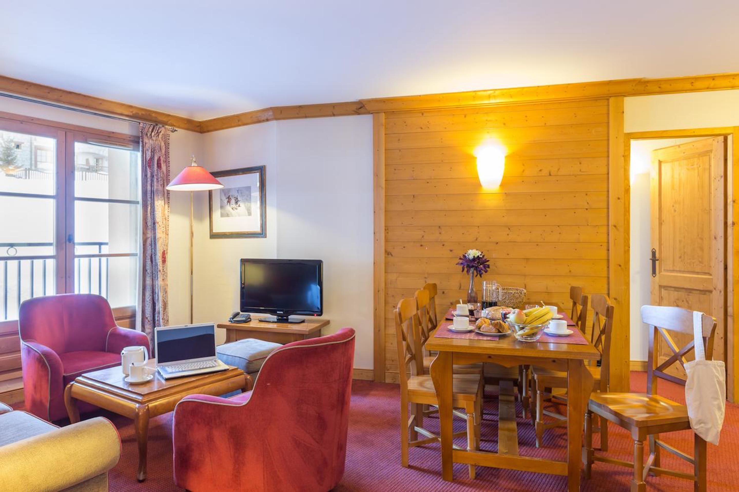 Une pièce chaleureuse avec un mur en bois, une table à manger entourée de chaises, un téléviseur et un ordinateur portable posé sur une petite table.