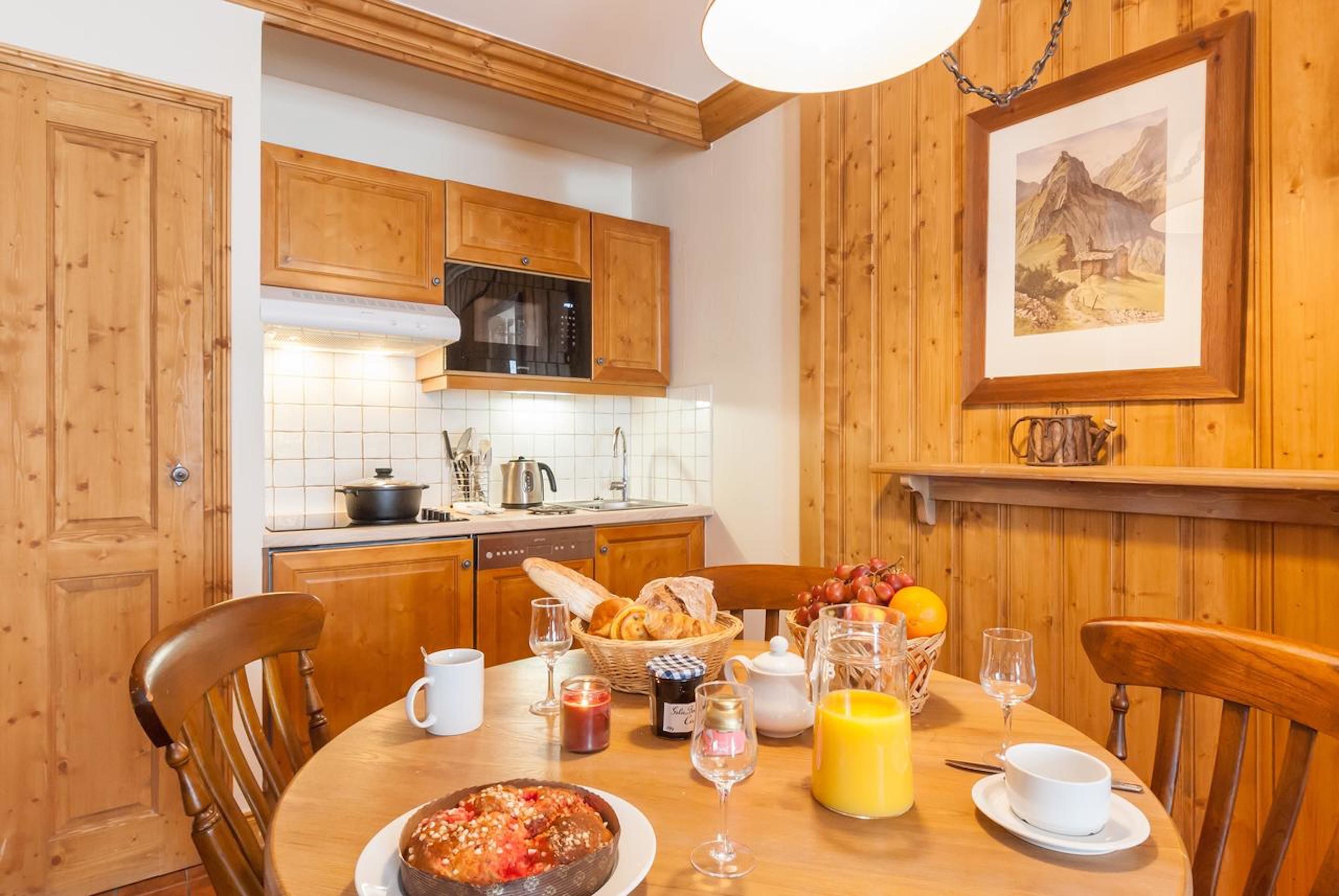 A wooden kitchen with a dining table set for breakfast, including bread, fruit, and a hot dish.