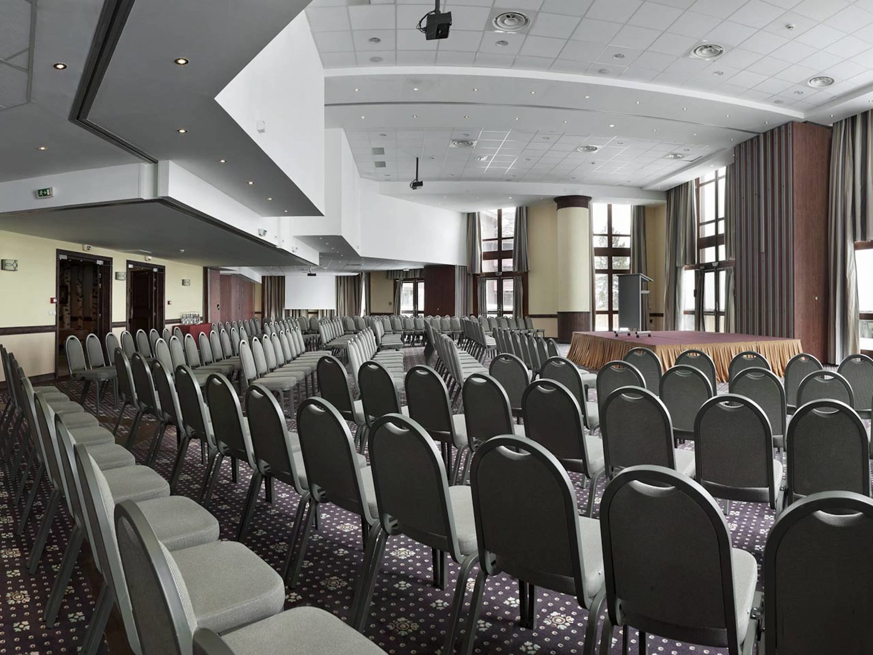 Uma sala de conferências com fileiras de cadeiras, um palco e janelas amplas.