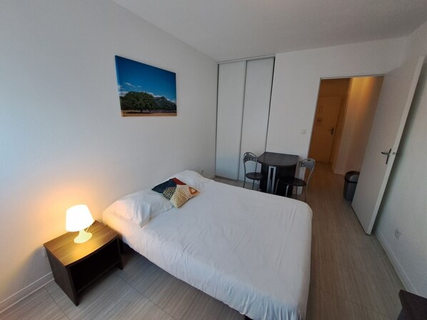 A bedroom with a bed, bedside table with a lamp, a small desk with chairs, a door, and a picture on the wall.