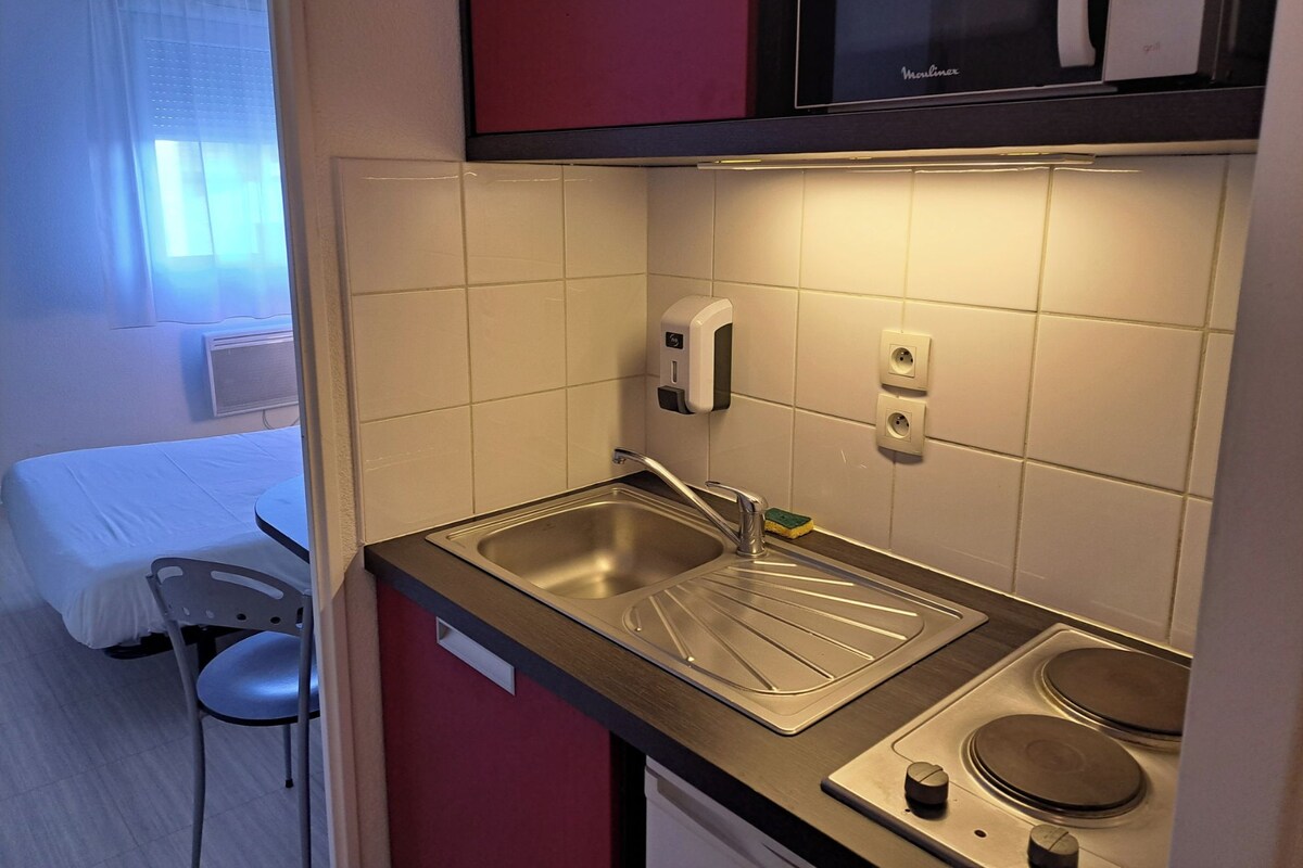 Una kitchenette compacta con un lavamanos, un microondas y una pequeña estufa. Se ve una cama y una silla en la habitación contigua.