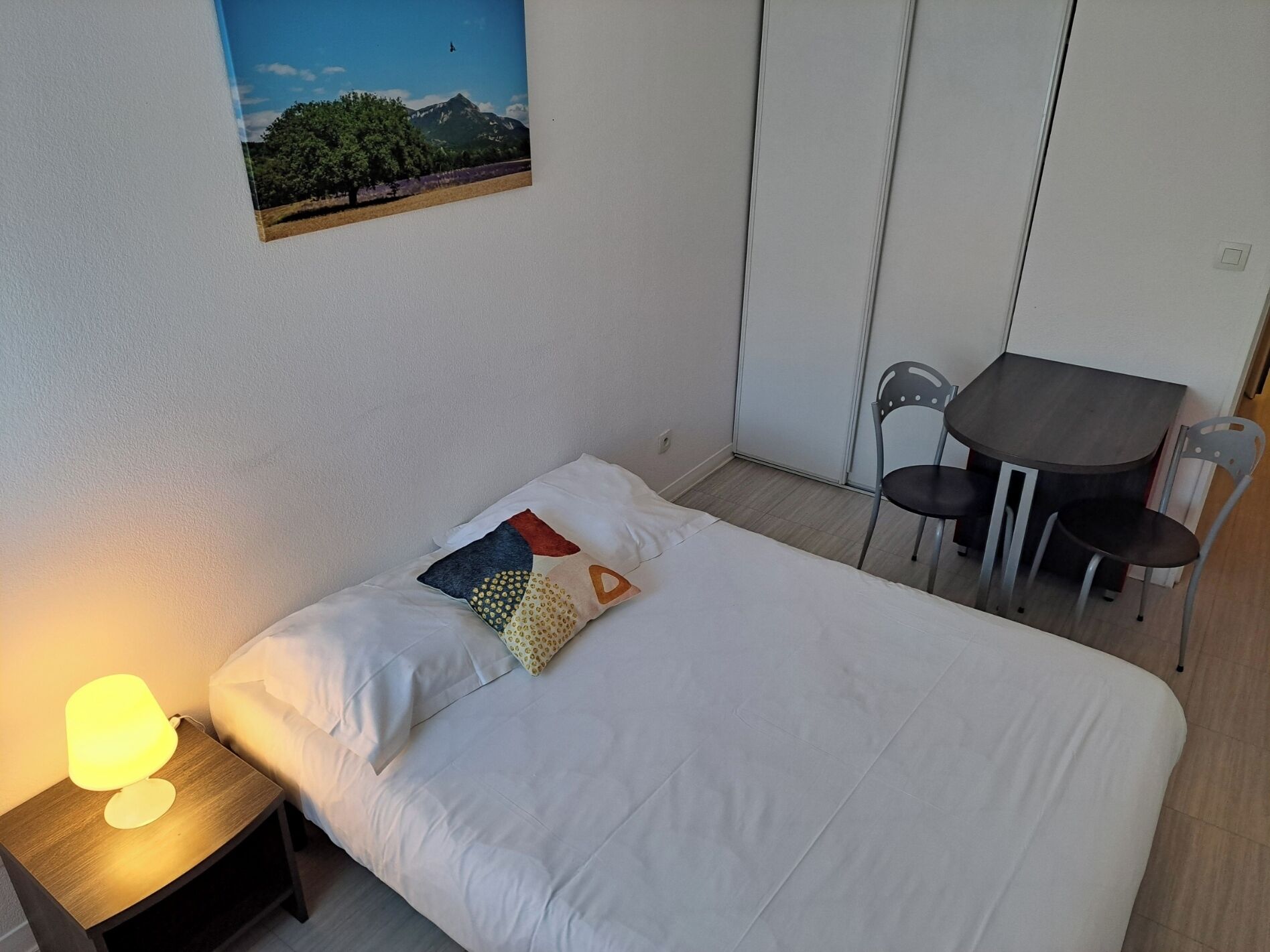 A single bed with white bedding, a bedside table with a lamp, and a small table with two chairs. A picture hangs on the wall above the bed.