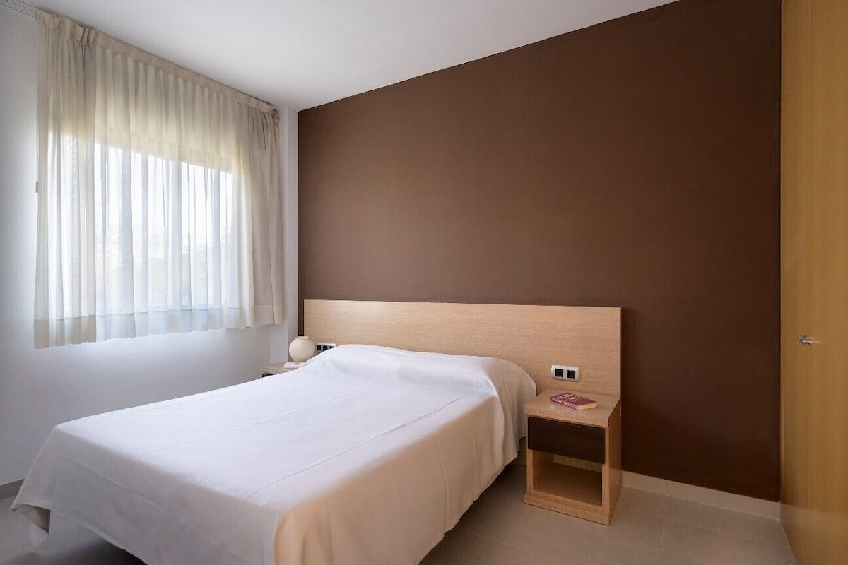 A hotel room with a single bed, a bedside table with a lamp, and a view of the outdoors through sheer curtains.