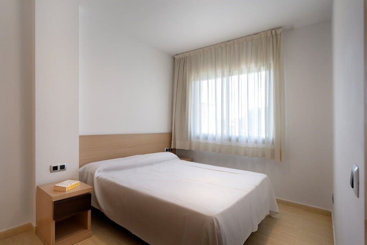 A hotel room with a single bed, a nightstand with a book, a window with sheer curtains, and a plain white wall.
