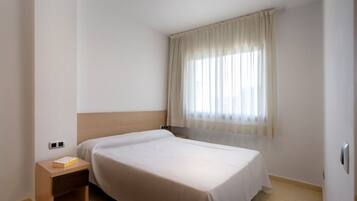 Habitación de hotel con una cama individual, una mesita de noche con un libro, una ventana con cortinas traslúcidas y una pared blanca lisa.