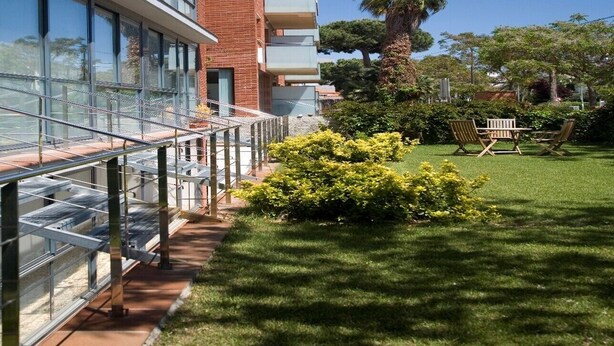 A modern building with a glass facade, a brick section, and a well-maintained lawn with outdoor furniture. - SG Costa BCN Apartments (Castelldefels)