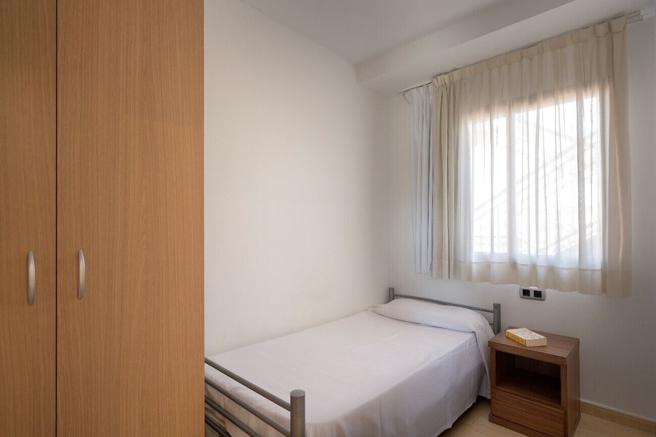 A small, single-bed room with a wooden wardrobe, a bedside table with a book, and a window with sheer curtains.