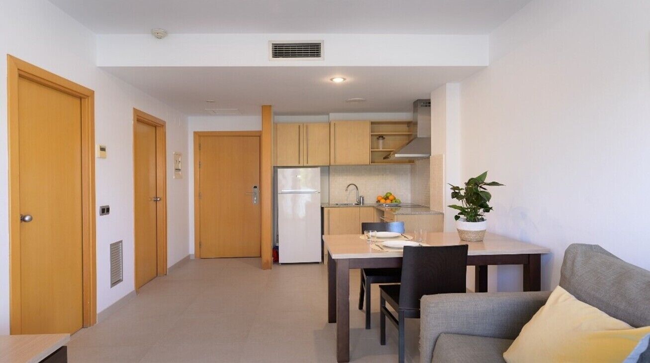 A compact kitchen with a refrigerator, sink, and wooden cabinets. There is a dining table with chairs and a potted plant.