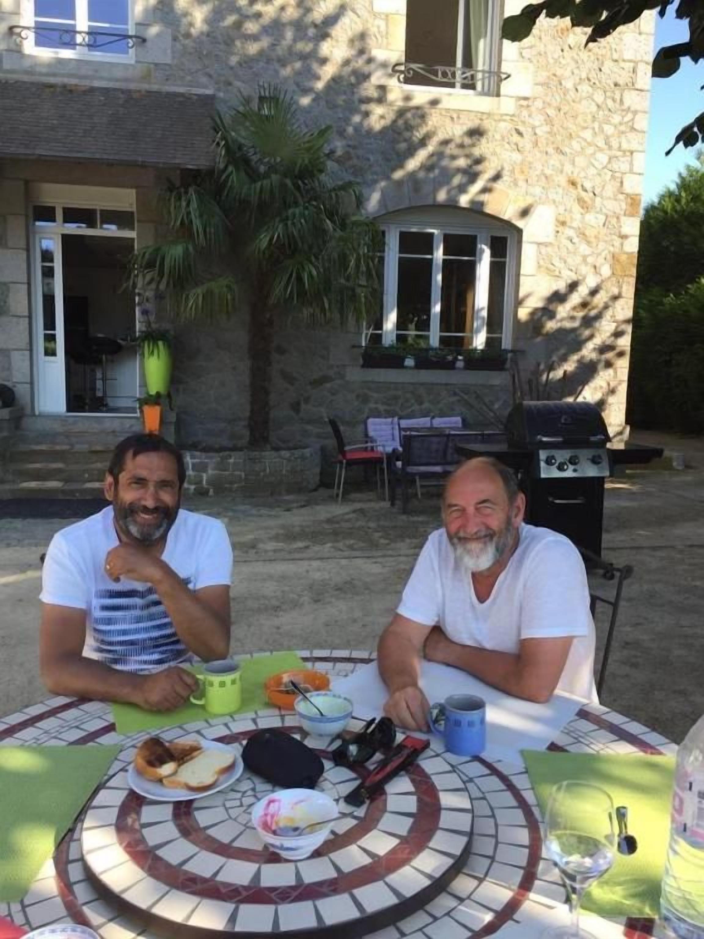 Dos personas sentadas en una mesa al aire libre con comida y bebidas, un edificio de piedra al fondo y una palmera.