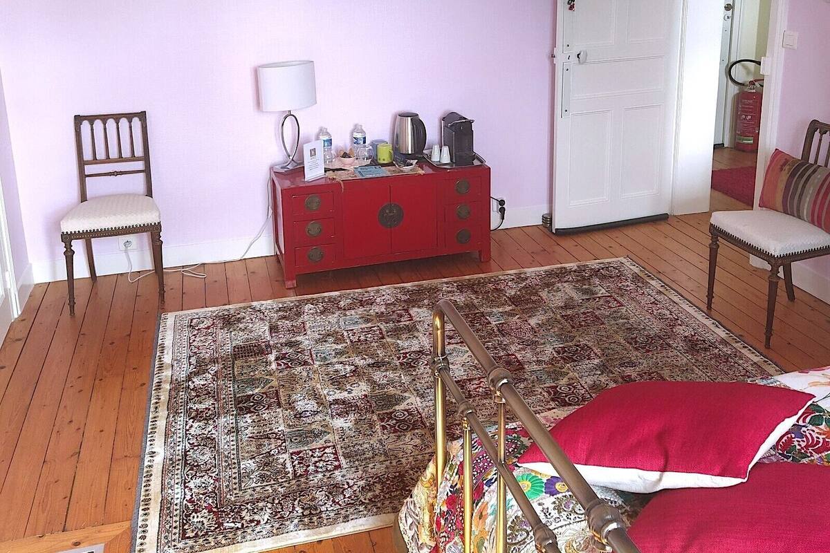 Une pièce avec un plancher en bois, une commode rouge, une chaise, un lit avec un oreiller rouge, un tableau encadré au mur et une porte blanche.