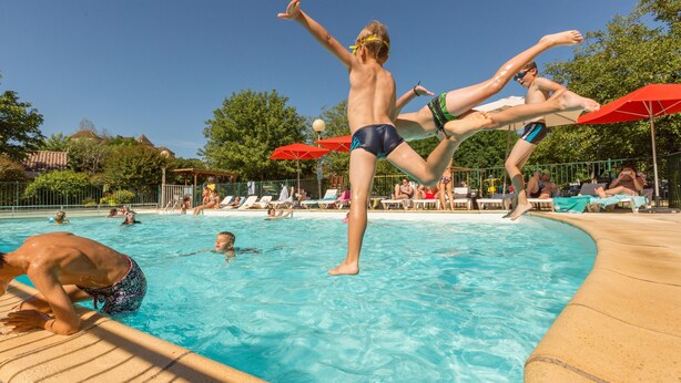 A swimming pool with people in the water and on the edge, surrounded by lounge chairs and umbrellas. - Camping la Plage (Meyronne)