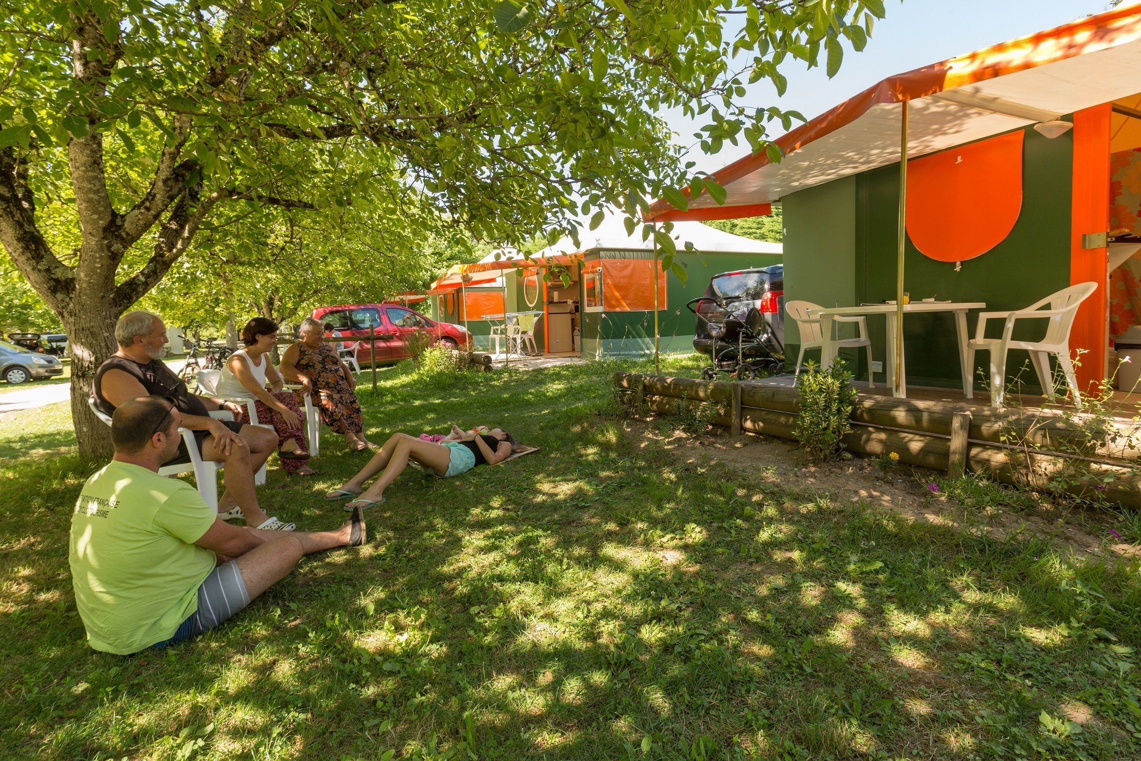 A group of people sitting on grass under a tree, with a green and orange building in the background.