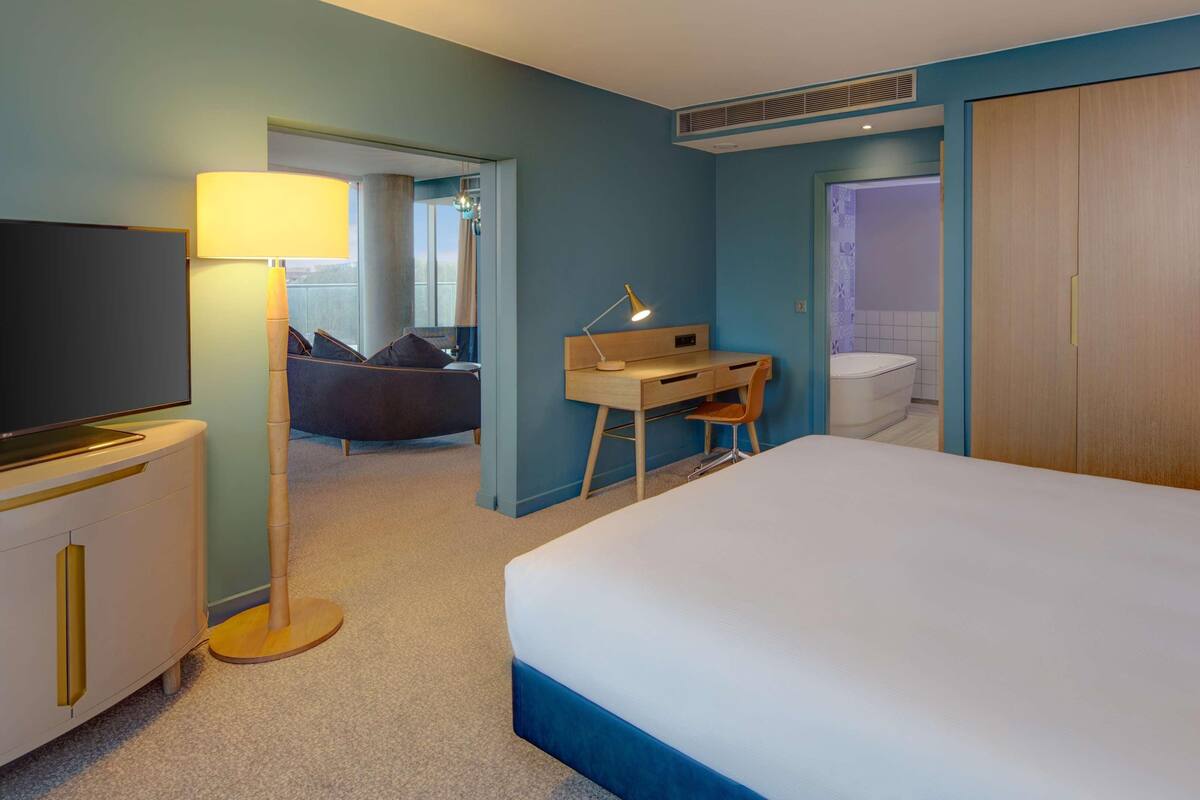 Ein Hotelzimmer mit einem großen Bett, einem Schreibtisch mit Stuhl, einem Fernseher und einem durch eine offene Tür einsehbarer Badezimmer.