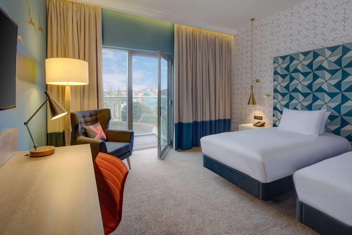 Ein Hotelzimmer mit einem großen Bett, einem Schreibtisch mit Lampe, einem Sessel und Blick auf eine Stadt durch eine Glasdoor.