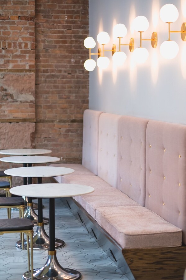 Une rangée de tables rondes avec des pieds en métal, un banc rembourré et un luminaire fixé au mur.
