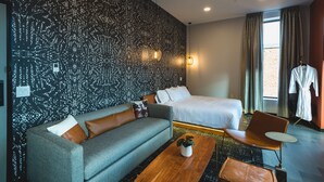 A modern hotel room with a patterned wall, a sofa, a bed, a chair, a small table, and a robe rack. - The Vault Hotel (Houghton)